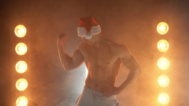 Victory gesture. Excited muscular guy in Santa hat showing win gesture, lamps illumination on background — Stock Video