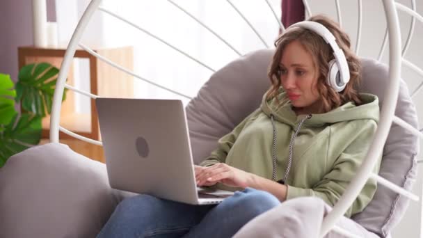 Feliz sonrisa mujer escuchar música auriculares uso portátil escribir mensaje de texto en el ordenador portátil mientras está sentado silla — Vídeos de Stock