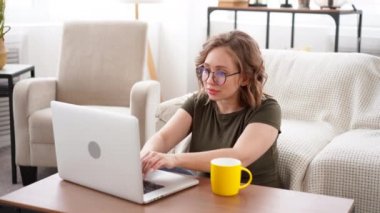 Gözlüklü kadın evde arka planda büyük pencereli koltukta otururken dizüstü bilgisayar kullanıyor. Görüntülü sohbet ev konsepti video konferansında çalışıyor. 