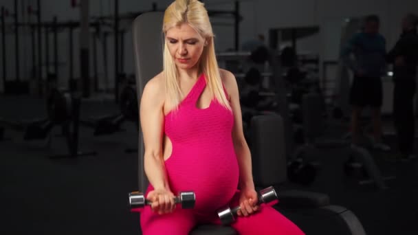 Ciężarna kobieta podnosząca hantle trening mięśni bicepsów na siłowni siedzi na ławce — Wideo stockowe