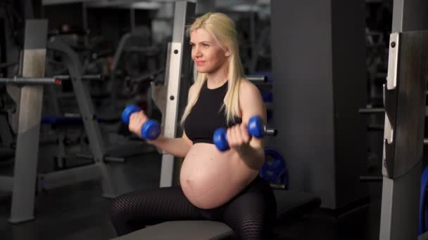 Беременная женщина поднимает гантели тренируя бицепсы мышцы в тренажерном зале сидя скамейка — стоковое видео