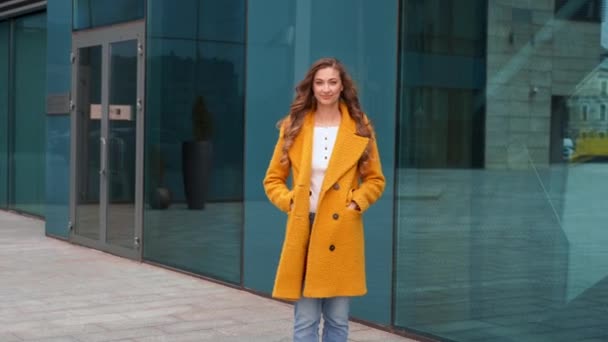 사업을 하는 여자 가 야외에 서 있는 노란 코트를 입고 있는 모습, 건물 뒷 배경에는 손을 흔들며 인사를 한다 — 비디오