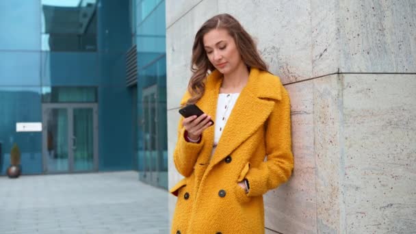 Donna d'affari vestita di giallo cappotto in piedi all'aperto edificio corporativo sfondo con smartphone digitando messaggio di testo Chiamare qualcuno — Video Stock