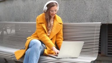 İş kadını gözlük takar müzik kulaklığı takar dışarıda oturur, dışarıda dizüstü bilgisayar kullanır.