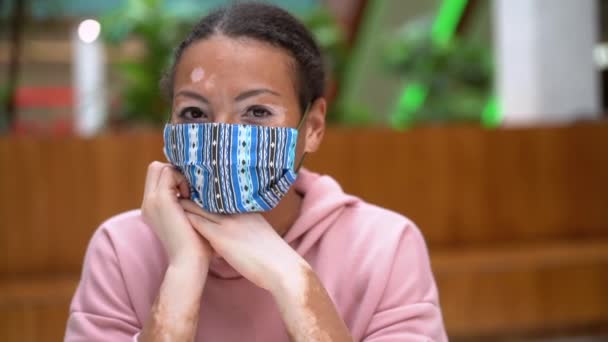 Mujer afroamericana negra con problema de pigmentación de la piel vitiligo con capucha rosa vestida en interiores y mascarilla protectora — Vídeo de stock