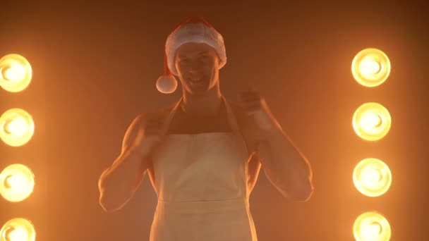 Горячий рождественский десерт. Мускулистый человек в фартуке и шляпе Санты показывает большой палец вверх жестом и улыбается в камеру, светильники освещения на заднем плане — стоковое видео