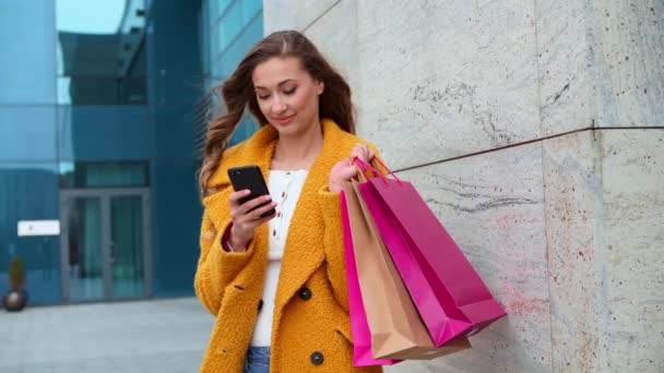 Mulher de negócios com sacos de compras vestido casaco amarelo andando ao ar livre corporativo edifício fundo uso smartphone compras on-line mensagem de texto — Vídeo de Stock