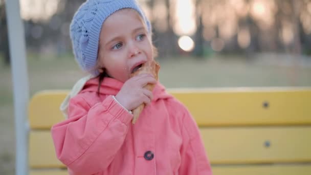 Маленькая девочка ест мороженое на открытом воздухе. Кавказская девочка наслаждается, облизывая белое мороженое в вафельном рожке — стоковое видео