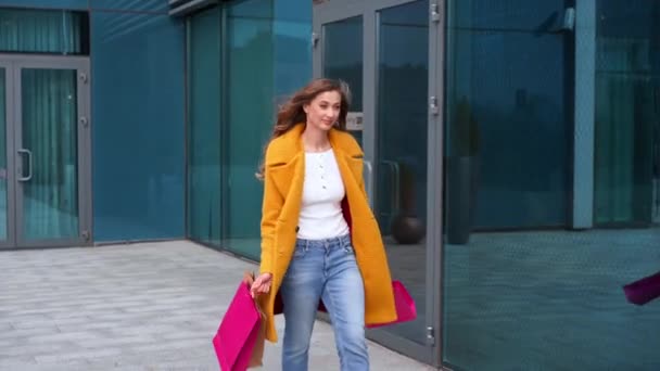 Femme d'affaires avec sacs à provisions habillé manteau jaune marchant à l'extérieur fond de bâtiment corporatif — Video