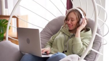 Neşeli gülümseyen kadın dinle müzik kulaklıkları laptopta mesaj yazarken koltukta oturuyor.