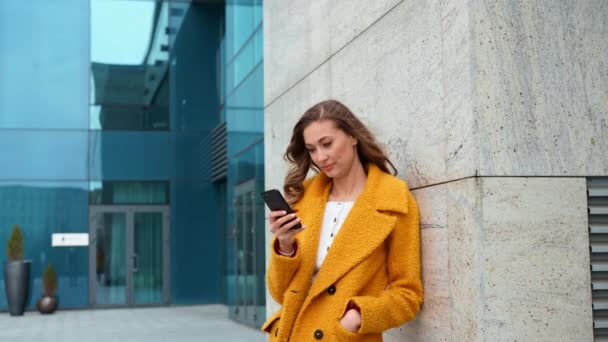 Mujer de negocios vestido abrigo amarillo de pie al aire libre edificio corporativo fondo con teléfono inteligente escribir mensaje de texto Llamar a alguien — Vídeos de Stock