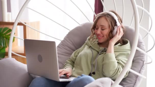Feliz sonrisa mujer escuchar música auriculares uso portátil escribir mensaje de texto en el ordenador portátil mientras está sentado silla — Vídeos de Stock