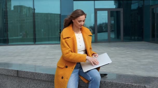 Geschäftsfrau mit Laptop gekleidet gelben Mantel sitzt draußen korporativen Gebäude Hintergrund — Stockvideo