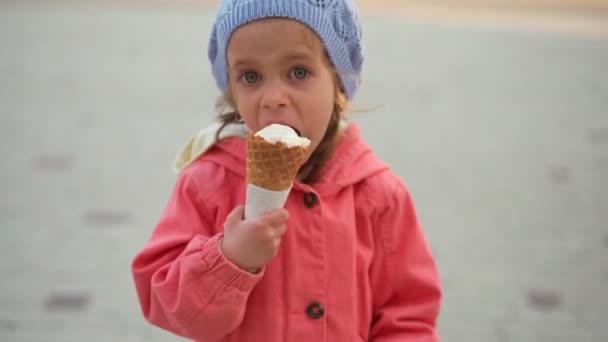 Маленькая девочка ест мороженое на открытом воздухе. Кавказская девочка наслаждается, облизывая белое мороженое в вафельном рожке — стоковое видео