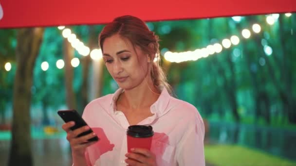 街の公園で雨の中で女性がキャノピーの下に立つ使用電話は手にコーヒーを保持しています — ストック動画