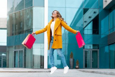 Alışveriş Çantalı İş Kadını Sarı Paltolu, Mutlulukla Atlayan Açık Hava Kurumsal Binası Kafkas Kadın İş Adamları Ofis Yakınındaki Şık İş Kadınları Sezon Satışı