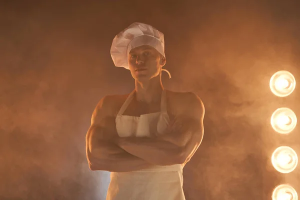 Portrait of muscular chef wearing white apron and chef hat, posing with folded arms on smoky background male housewife. husband in kitchen. brutal butcher.