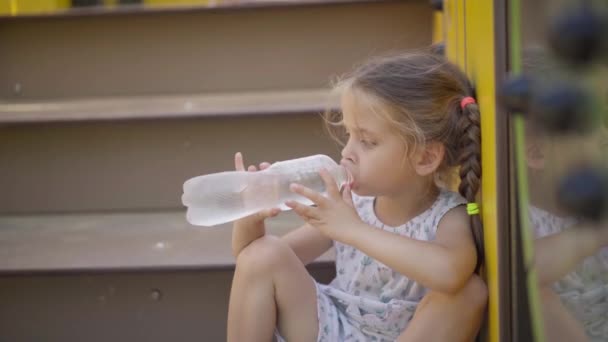 Petite fille boire de l'eau bouteille en plastique au repos après jeu actif sur aire de jeux journée d'été — Video