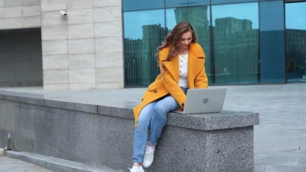 Mujer de negocios usando portátil vestido abrigo amarillo sentado al aire libre corporativo edificio de fondo — Vídeos de Stock