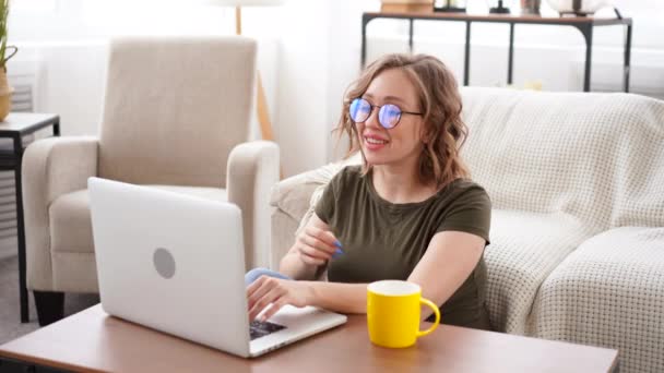 Жінка в окулярах використовує портативний комп'ютер, сидячи на дивані з великим вікном на фоні домашнього інтер'єру розмовного відео чату Робота вдома концепція Відеоконференція — стокове відео