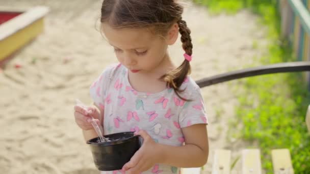 Mała dziewczynka jeść lody łyżka odkryty plac zabaw plastikowa płyta Letni dzień slow motion — Wideo stockowe