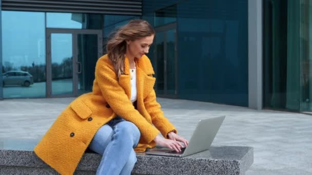 Biznes kobieta za pomocą laptopa ubrany żółty płaszcz siedzi na zewnątrz korporacyjne tło budynku — Wideo stockowe