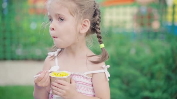 Маленькая девочка ест кукурузную бумагу стекло сидя скамейка летний парк — стоковое видео
