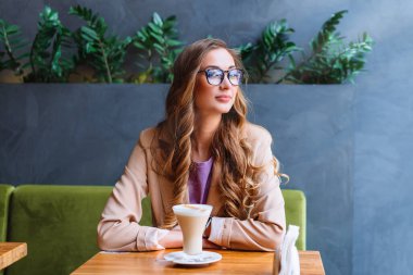 İş kadını, Pencere kenarında oturan, iş ortağını bekleyen beyaz kadın, dinlenme kafeteryası, geç saatte kahve içen, dışarı bakan, gülümseyen kadın.