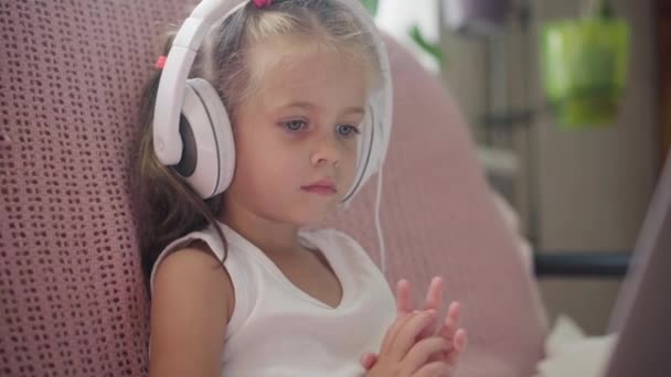 Mała dziewczynka siedzi sofa w słuchawkach Słuchać muzyki Użyj laptopa Baw się dobrze Strona główna Wnętrze Kaukaski Kobieta Dziecko Użyj technologii Nowoczesne dzieci Szczęśliwe dzieciństwo — Wideo stockowe