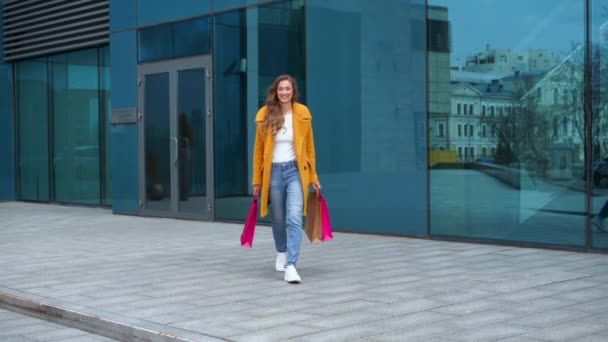 Alışveriş çantalı bir iş kadını sarı ceketli dışarıda yürüyen kurumsal bina arka planında dans eden Mutlu Komik — Stok video