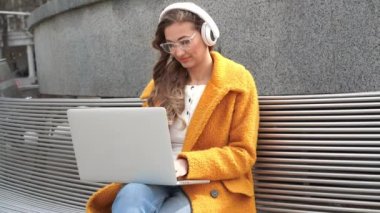 İş kadını gözlük takar müzik kulaklığı takar dışarıda oturur, dışarıda dizüstü bilgisayar kullanır.