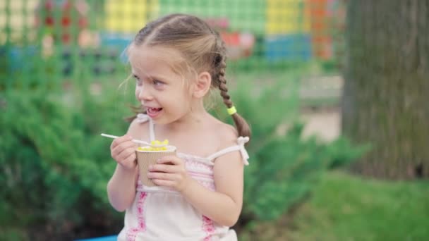 Malá holka jíst kukuřičný papír sklo sedací lavice letní park — Stock video