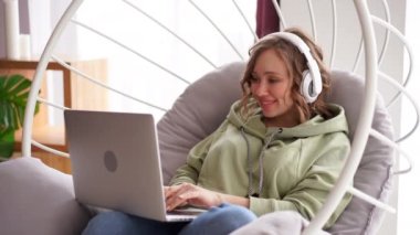 Neşeli gülümseyen kadın dinle müzik kulaklıkları laptopta mesaj yazarken koltukta oturuyor.