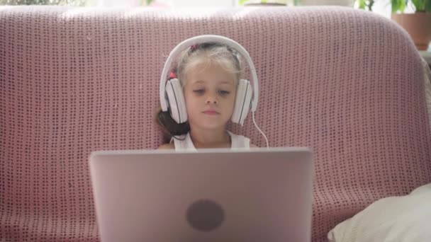 Маленька дівчинка сидить на дивані в навушниках Слухайте музику Використовуйте ноутбук Розважайтеся домашнім інтер'єром Кавказька жінка Технологія використання дітей Сучасні діти Щасливе дитинство — стокове відео