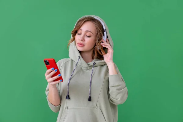 Happy teeth smile woman listen music headphones holding smartphone in hand looking screen Caucasian female enjoy podcast or audio books dressed oversize hoodie green background close up portrait