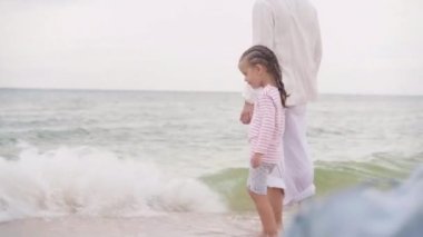 Anne, küçük kızıyla birlikte sahilde mutlu bir ailede yürüyor. Anne, deniz kıyısında küçük bir çocuğun elini tutuyor. Seyahat Yaz Tatili 