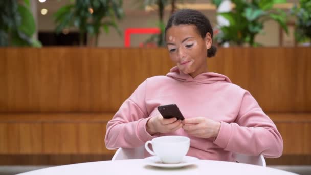 Чорно-афроамериканська жінка з вітиліго проблеми зі шкірою в приміщенні одягнена рожева капуста використовує смартфон. — стокове відео