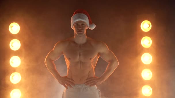 Portrait de l'homme musclé portant le chapeau de Noël Père Noël, montrant le doigt sur la caméra sur fond fumé — Video