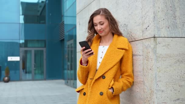 ビジネス女性は、スマートフォンの入力テキストメッセージと屋外の企業の建物の背景に立って黄色のコートを着て誰かを呼び出します — ストック動画
