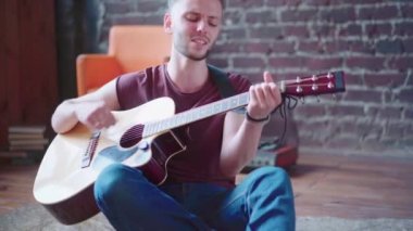 Akustik gitar çalan yakışıklı genç adam oturma odasında oturuyor.