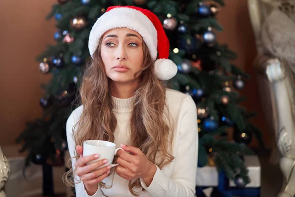 Christmas Woman dressed white sweater Santa hat sitting floor near christmas tree  Caucasian female relaxing winter holiday home interior drink hot morning coffee and upset. It was a bad year Boring