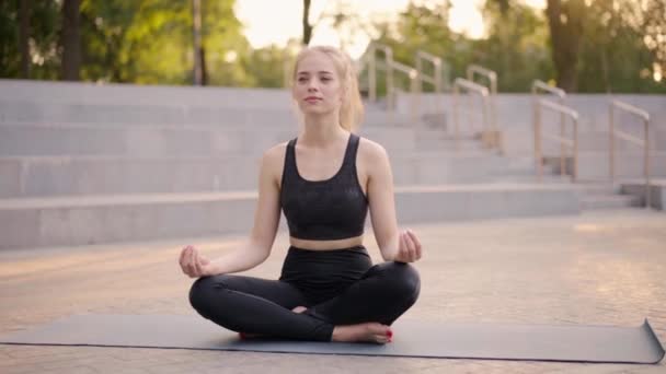 Yoga e meditação na cidade moderna Mulher caucasiana Relaxe posição de lótus Esteira de ioga sentada ao ar livre Parque de verão no chão de concreto — Vídeo de Stock