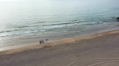 Aerial drone shot capturing sandy beach with surfers in sea. The waves gently break along the shore and group of people gathered on beach enjoying holiday