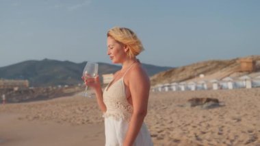 Beyaz elbiseli genç bir kadın kumlu sahilde şarap içiyor okyanus dalgaları ve hafif bir esintiyle huzurlu bir sahil hayatı sürüyor ve yaz tatilinin tadını çıkarıyor.