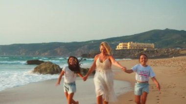 Mutlu anne, gün batımında kumlu sahilde kızlarıyla el ele tutuşup neşeli aile bağlarının ve deniz kenarında neşeli tatilin tadını çıkarıyor.