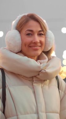Rahat bir kışlık palto giyen neşeli bir kadın ve tüylü kulaklıklar ışıl ışıl bir kış ortamında ışıl ışıl tatil ışıkları arasında ışıl ışıl gülümsüyor. Noel ruhunu somutlaştırıyor..