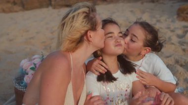 Anne ve kızı, kumsalda genç bir kızı yanaklarından öpüyor. Yaz tatilinde sevgi, şefkat ve aile bağlarını gösteriyor.
