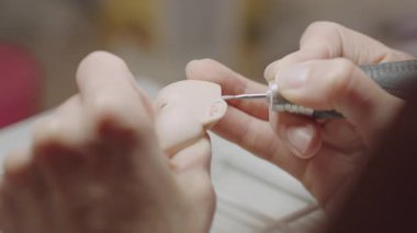 Oyuncak bebek kafasının kulak detaylarını oymak için el bilgisayarı kullanan sanatçıya yakın çekim, bebek heykel sürecinde hassasiyet ve gerçekçiliğe odaklanma