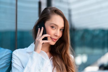 Modern ofis binasının dışında akıllı telefondan konuşurken uzun kahverengi saçlı, gülümseyen ve kameraya bakan bir iş kadını. Güven ve profesyonellik yayıyor.