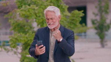 Akıllı telefona bakarken heyecanla yumruk atan neşeli, olgun iş adamı zaferi kutluyor ya da ofis binasının dışında neşeli bir ifadeyle başarıyı kutluyor.